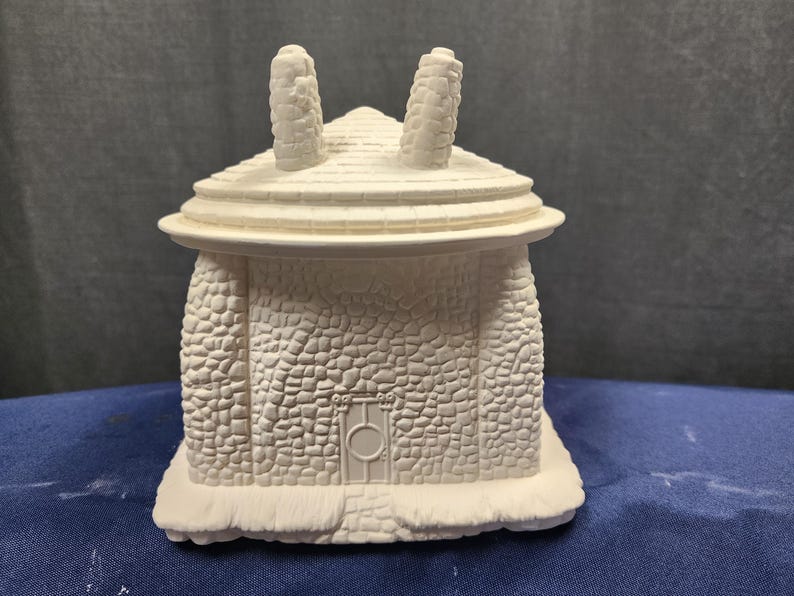 May include: White ceramic house figurine with a stone-like texture and a small door. The house has two chimneys and a rounded roof.