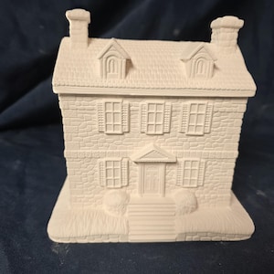 May include: A white ceramic house with a textured stone facade, two chimneys, and a detailed roof. The house features windows with shutters and a front door with steps, perfect for painting or decorating.