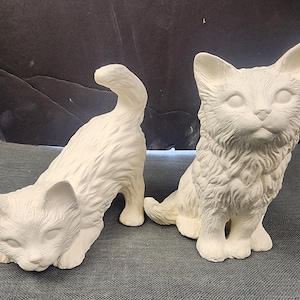 May include: Two white ceramic cat figurines. One cat is sitting with its tail curled around its legs. The other cat is standing with its head down and its tail raised.