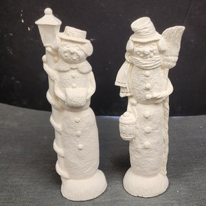 May include: Two white ceramic snowman figurines. One snowman is holding a lantern and the other is holding a bucket. Both snowmen are wearing hats and scarves.