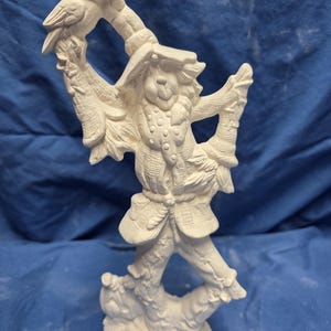 May include: A white ceramic figurine of a scarecrow holding a branch with a bird perched on it. The scarecrow wears a hat, jacket, and pants, all with detailed textures. The figurine is unpainted and stands on a base.