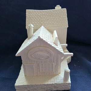 May include: A white ceramic miniature of a fishing shop with a fish emblem above the door. The shop has a textured roof, chimney, and a small dock. The base is made to look like stone.