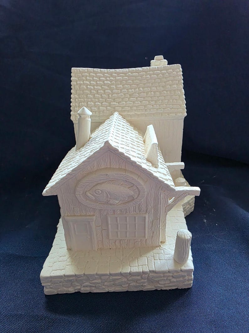 May include: A white ceramic miniature of a fishing shop with a fish emblem above the door. The shop has a textured roof, chimney, and a small dock. The base is made to look like stone.