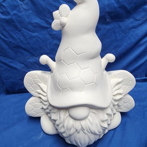 May include: A white ceramic gnome figurine with a long, curved hat, a textured beard, and bee-shaped wings. The hat features a honeycomb pattern. A flower is attached to the hat. The figurine is unpainted and ready for customization.
