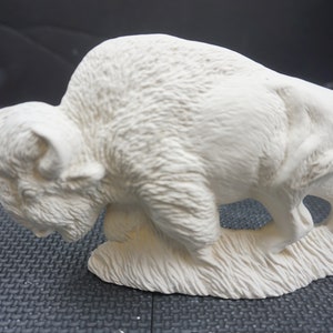 May include: White ceramic bison sculpture with detailed fur texture. The bison is in a walking pose with its head lowered. The sculpture is on a base with a textured grass design.