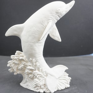May include: A white dolphin sculpture leaping from a wave. The dolphin is sculpted with detailed features, including fins, a tail, and facial details. The sculpture is made of a solid, opaque material.