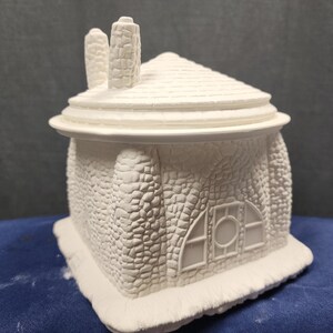 May include: White ceramic cottage-style house with a stone-like texture and a window with a round center. The house has a roof with two chimneys.
