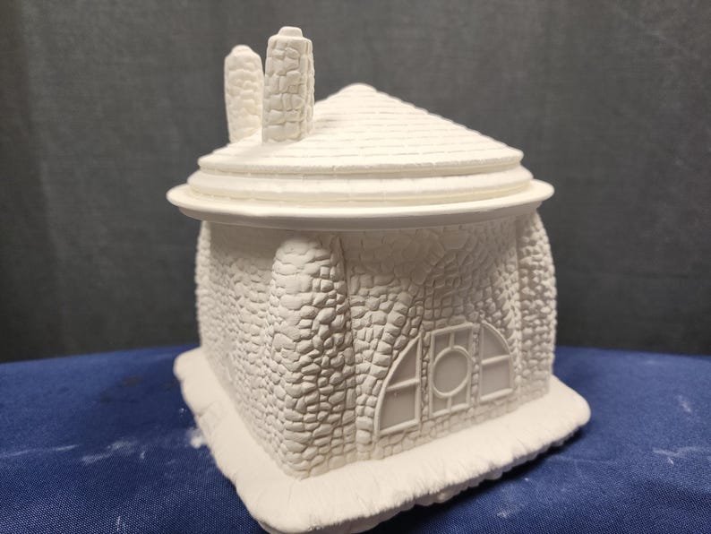 May include: White ceramic cottage-style house with a stone-like texture and a window with a round center. The house has a roof with two chimneys.
