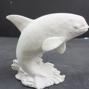 May include: White ceramic figurine of an orca whale leaping out of the water. The orca is detailed with fins and a dorsal fin. The base of the figurine is textured to resemble waves.