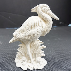 May include: A cream-colored ceramic bird figurine, possibly a heron or egret, perched on a stylized base of reeds. The sculpture has detailed feather textures and a long beak. The bird is a decorative home accent.