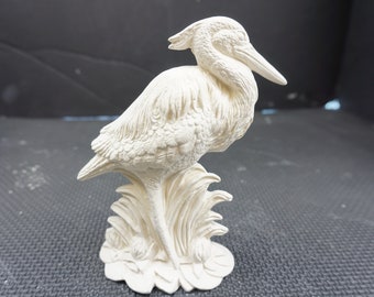 May include: A cream-colored ceramic bird figurine, possibly a heron or egret, perched on a stylized base of reeds. The sculpture has detailed feather textures and a long beak. The bird is a decorative home accent.