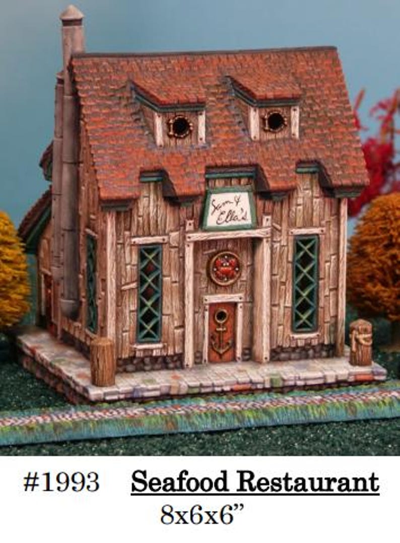 Puede incluir: Un restaurante de mariscos en miniatura con un exterior de madera desgastada y un tejado de tejas rojas. El edificio presenta un letrero que dice "Sam & Ella's" y mide aproximadamente 20x15x15 cm. La puerta principal tiene un dise&ntilde;o de ancla.