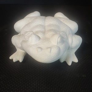 May include: White ceramic frog figurine with a detailed textured surface and large eyes. The frog is in a sitting position with its legs outstretched.