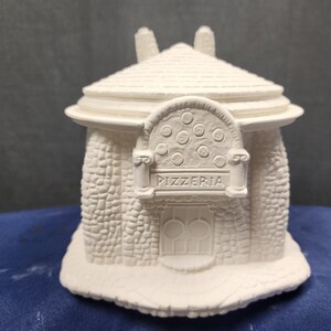 May include: White ceramic figurine of a pizzeria building with a rounded roof, stone facade, and a sign that reads "Pizzeria".