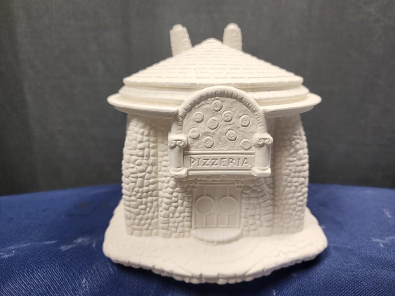 May include: White ceramic figurine of a pizzeria building with a rounded roof, stone facade, and a sign that reads "Pizzeria".