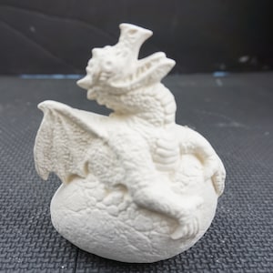 May include: White ceramic dragon figurine sitting on a white ceramic egg. The dragon has wings and a spiky crest.