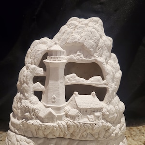 May include: White ceramic lighthouse figurine with a small house and rocky cliffs. The lighthouse has a detailed design and is set against a backdrop of a cave.