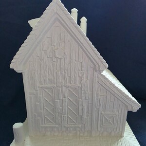 Puede incluir: Una caba&ntilde;a en miniatura de cer&aacute;mica blanca con un exterior texturizado similar a la madera. La caba&ntilde;a presenta un tejado a dos aguas, dos chimeneas y dos ventanas con patrones entrecruzados. Tambi&eacute;n son visibles un peque&ntilde;o porche y un elemento decorativo.