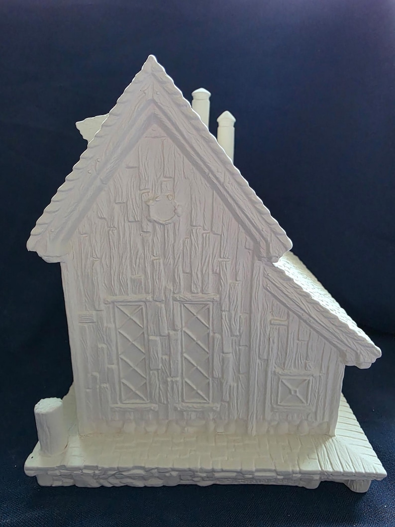 Puede incluir: Una caba&ntilde;a en miniatura de cer&aacute;mica blanca con un exterior texturizado similar a la madera. La caba&ntilde;a presenta un tejado a dos aguas, dos chimeneas y dos ventanas con patrones entrecruzados. Tambi&eacute;n son visibles un peque&ntilde;o porche y un elemento decorativo.