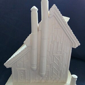 Puede incluir: Figurita de casa de cer&aacute;mica de color crema con dos chimeneas altas y una chimenea peque&ntilde;a en la base. La casa tiene un tejado y un revestimiento texturizados, de estilo teja, y una ventana decorativa.