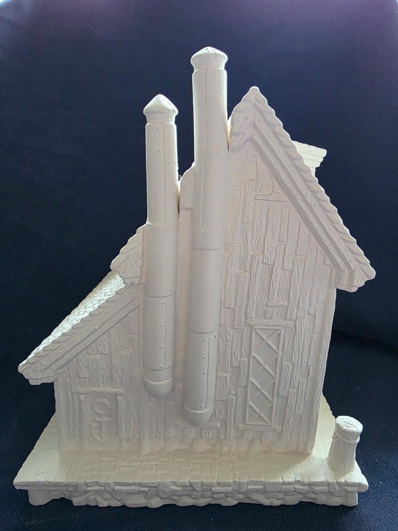 Puede incluir: Figurita de casa de cer&aacute;mica de color crema con dos chimeneas altas y una chimenea peque&ntilde;a en la base. La casa tiene un tejado y un revestimiento texturizados, de estilo teja, y una ventana decorativa.