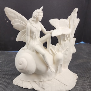 May include: White ceramic figurine of a fairy sitting on a snail with a butterfly on a flower. The fairy has wings and is wearing a dress. The snail is on a base with a flower.