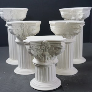 May include: Set of five white ceramic pedestal columns with ornate details. The columns are arranged in a staggered formation, with the largest column in the center and the smallest columns on either side.