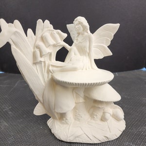 May include: A white ceramic fairy figurine. The fairy is seated on a mushroom, with wings spread. The figurine is surrounded by sculpted leaves and flowers, creating a whimsical, fantasy-themed decorative piece.