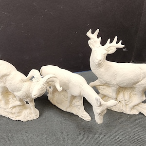May include: Three white ceramic animal figurines, a deer, a ram, and a mountain goat, are posed on a gray surface. The deer is standing on a rock with its head turned to the side. The ram and mountain goat are both standing on rocks with their heads down.