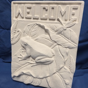 May include: A rectangular, off-white welcome sign with a frog, dragonfly, and leaf design. The word "WELCOME" is embossed at the top. The sign features a detailed relief of a frog resting on large leaves, with a dragonfly nearby.