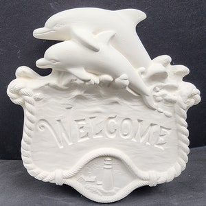 May include: White ceramic welcome sign with two dolphins jumping over waves. The sign has a rope border and the word "WELCOME" is embossed on the front. The bottom of the sign has a lighthouse and a small building.