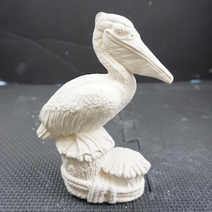 May include: White ceramic figurine of a pelican perched on a wooden post. The pelican has a detailed beak and feathers.