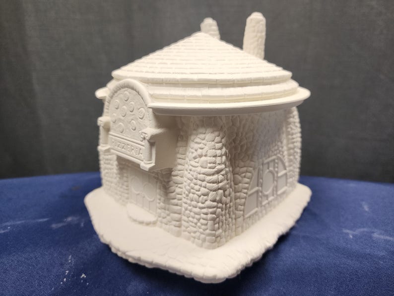 May include: White ceramic model of a small building with a stone facade and a sign that reads "Pizzeria".