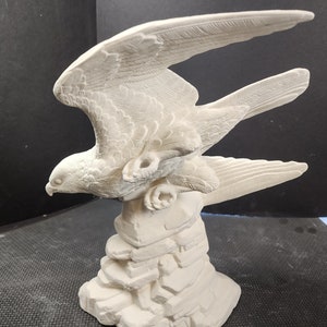 May include: A detailed, off-white sculpture of a bird of prey in flight. The bird is perched atop a rocky base, with wings outstretched, showcasing intricate feather detailing. The sculpture is made of a solid, possibly ceramic, material.
