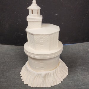 May include: White ceramic lighthouse figurine with a detailed design. The lighthouse has a round base with a textured surface, a cylindrical tower with a ladder, and a square top with a lantern.