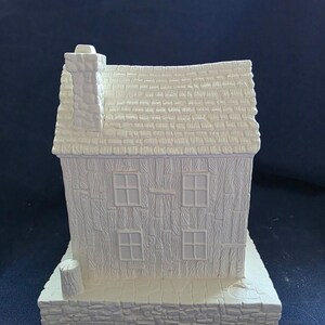 May include: A white ceramic house figurine with a textured roof, four windows, and a chimney. The house sits on a stone-like base. The overall design is a rustic, cottage-style theme.