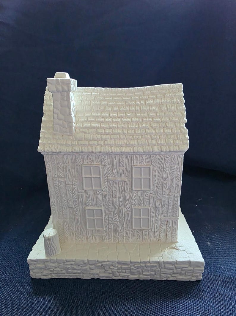 May include: A white ceramic house figurine with a textured roof, four windows, and a chimney. The house sits on a stone-like base. The overall design is a rustic, cottage-style theme.