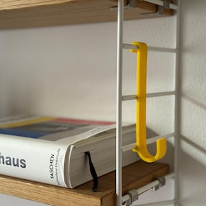Peut inclure: Un crochet jaune vif suspendu à une étagère métallique blanche. Un livre avec le mot "bauhaus" est posé sur l'étagère en bois en dessous. Le système d'étagères est fixé au mur.
