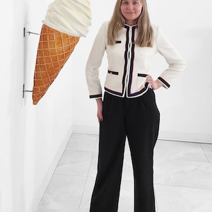 May include: A woman wearing a white jacket with black and red trim and black pants stands in front of a white wall with a large, white and brown ice cream cone hanging on the wall.