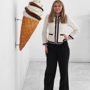 May include: A woman wearing a white jacket with black and red trim stands in front of a large, white and brown ice cream cone.