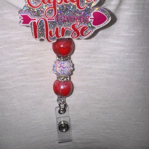 May include: A silver glitter badge reel with the text "Cupid's Favorite Nurse" and a pink arrow and heart. The reel has three red beads with white glitter and rhinestones.