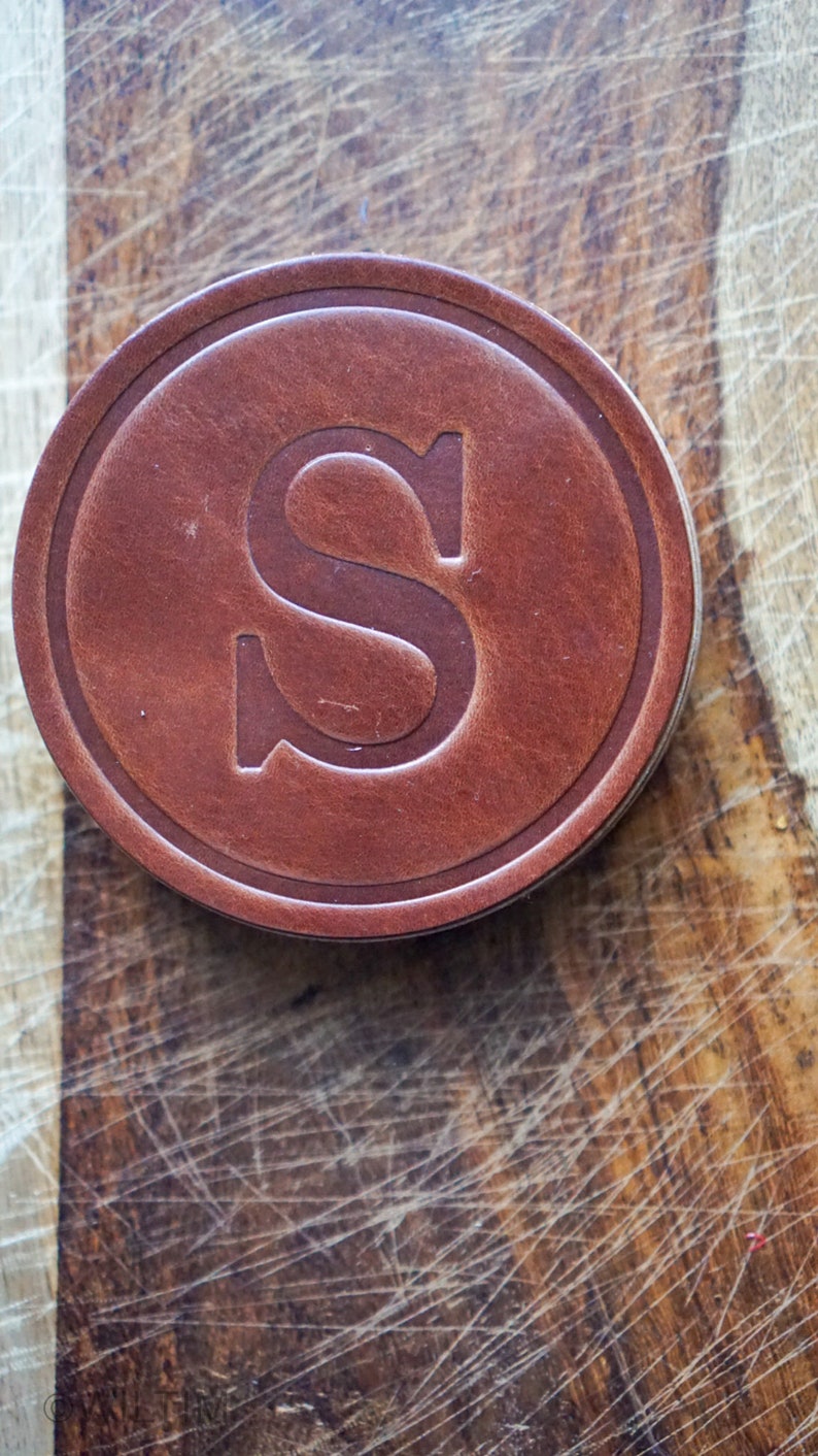 May include: A circular, brown leather coaster featuring a debossed letter "S". The coaster has a raised border and sits on a wooden surface with visible scratches.