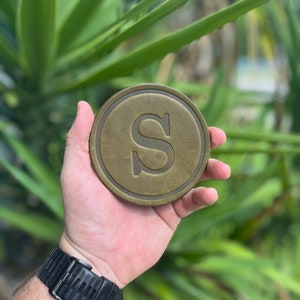 May include: A circular, bronze-toned coaster featuring a raised edge and the letter "S" in the centre. The coaster is held in a hand, with a black watch visible on the wrist. The background shows blurred green foliage.