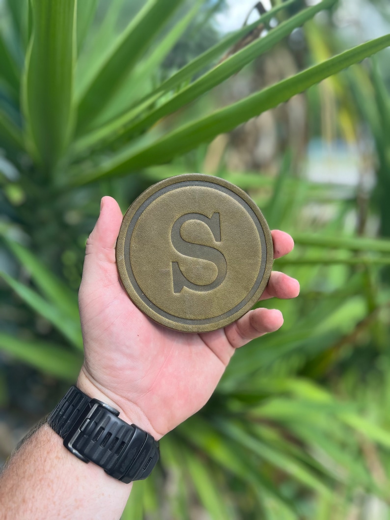 May include: A circular, bronze-toned coaster featuring a raised edge and the letter "S" in the centre. The coaster is held in a hand, with a black watch visible on the wrist. The background shows blurred green foliage.