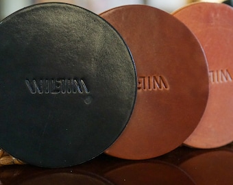 May include: Three round leather coasters with the word "WILKIN" embossed on each. The coasters are black, brown, and a reddish-brown colour.