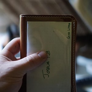 Puede incluir: Una cartera de cuero marrón con un inserto de tarjeta blanco. El inserto de tarjeta tiene el texto "Joshua 1:3" y "Good Word" escrito con tinta verde.