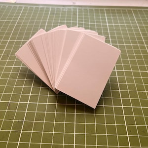 May include: A stack of light beige rectangular cards fanned out on a green cutting mat. The cards are neatly arranged, with their edges visible, creating a layered effect. The background is a grid pattern.