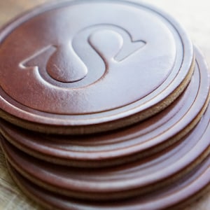 May include: A pile of brown leather coasters with a debossed design. The coasters are circular with a lighter brown edge and a raised, stylized letter "S" in the centre. The coasters are stacked on a wooden surface.