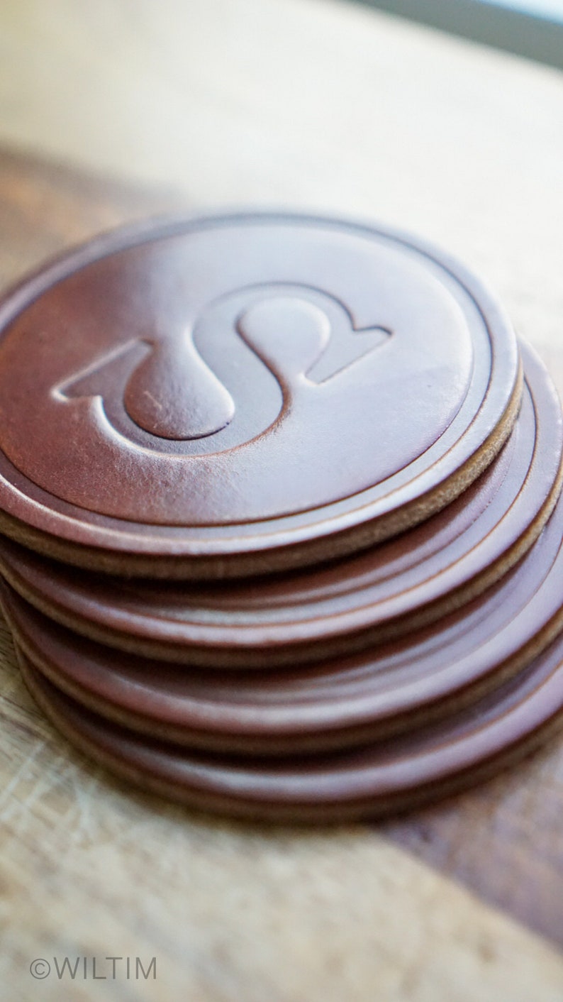 May include: A pile of brown leather coasters with a debossed design. The coasters are circular with a lighter brown edge and a raised, stylized letter "S" in the centre. The coasters are stacked on a wooden surface.