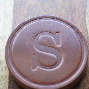 May include: A brown, circular coaster with a raised edge and a large, embossed letter "S" in the centre. The coaster is made of a solid material and rests on a wooden surface.